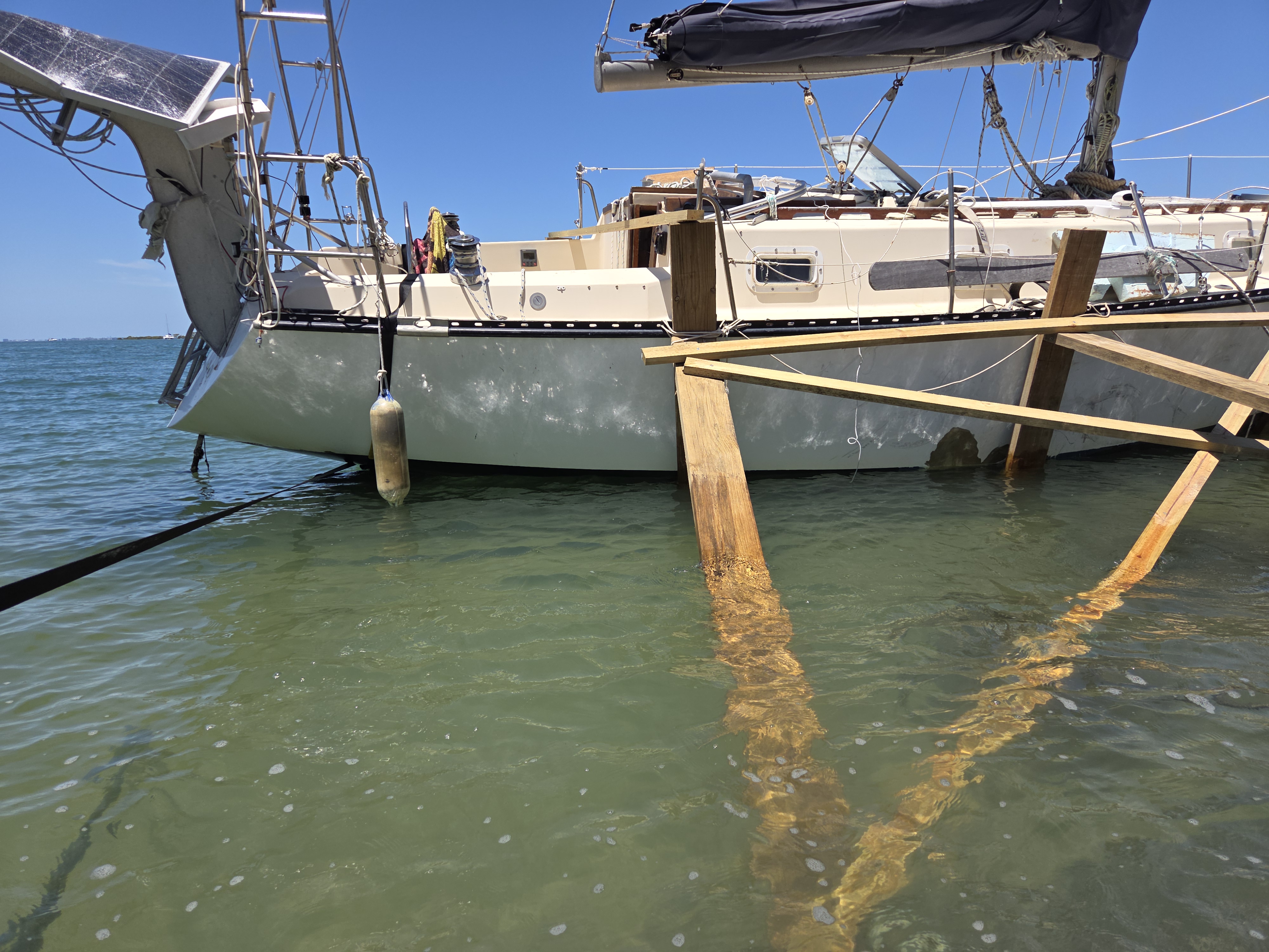 Sailing vessel back on the water during repair, supported by a temporary timber cradle, clear blue sky